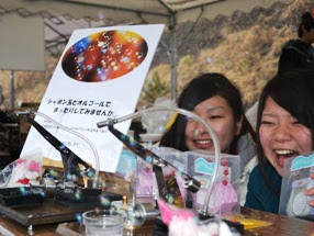 屋外のテントの下で、若い女性二人が笑顔で写り、目の前の机にはビーズや道具、完成したアクセサリーが並べられている写真