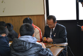 生徒と一緒になってまゆ細工に取り組んでいるスーツ姿の男性の写真