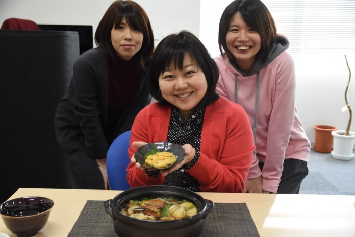 たくさんの具が入った土鍋が置かれたテーブルを前に、椅子に座ったリポーターの女性がホヤが入った小皿を両手で持ち、その後ろに2人の女性が笑顔で立っている写真