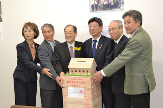 屋根付きの小さな家の形をした「書き損じはがき回収ポスト」が中央に置かれ、その後ろで佐藤町長と関係者の方々男女6人がポストに手を添え、笑顔でカメラに向かって並んでいる記念写真