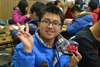 完成した赤い花のコサージュが入った透明なケースを手に、笑顔でピースサインをしている男子生徒の写真