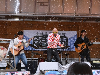 雪が散る中、イベント会場の舞台で赤い水玉模様のシャツを着たサンプラザ中野くんが中央でマイクを持って歌い、左右にギターを弾く男性二人が立ちライブイベントが行われている写真