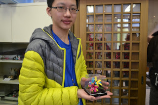 黄色、水色、ピンクなどの繭を使ったカラフルな花のコサージュが入った透明なケースを両手で持ってこちらに見せている男子生徒の写真