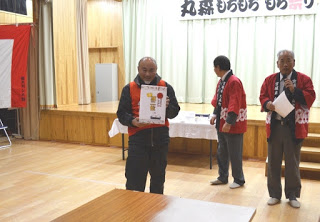 赤い法被を着てマイクを持った男性と同じ法被の男性が立つ横で、2位の賞品の箱を胸の前で持ち笑顔の男性の写真