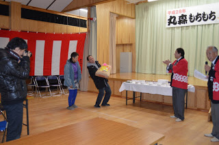 ステージ前に置かれた白いクロスが掛かったテーブルの右側に、赤い法被を着てマイクを持った男性と同じ法被の男性が立ち、左側にはグランプリ賞品の米袋を重たそうに抱える男性と女性が並んで立っている場面をカメラを持った男性が撮影している様子の写真