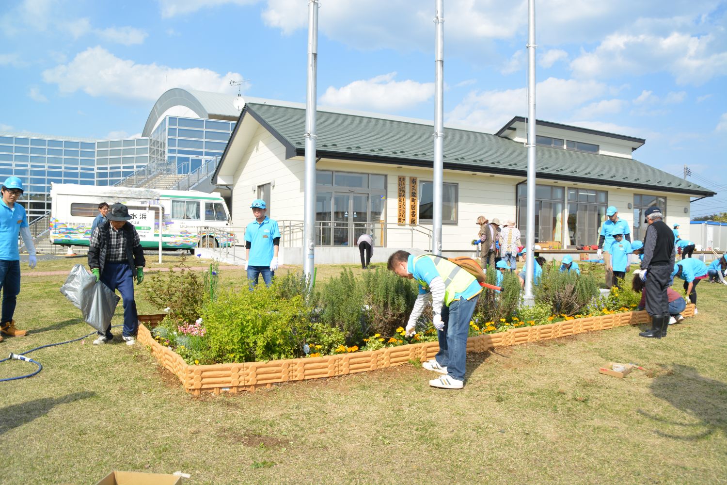 お揃いの水色のキャップにシャツを着た多くの人々が、花壇の手入れや清掃活動をしている様子の写真