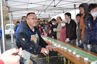 テントの下に並べられた長机の前に多くの参加者が並び、机の上にはラップに包まれたごはんが並べられていて、20メートルののり巻きを作る準備が進んでいる様子の写真