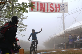 霧に包まれたゴール地点で、「FINISH」と書かれた横断幕の下を、マウンテンバイクに乗った選手が片手を挙げてゴールしている写真