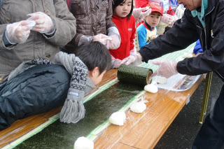 ラップに包まれたごはんが長いテーブルに並べられ、その横にはくるまれた海苔が敷かれていて、子どもが海苔に顔を近づけてみている様子の写真