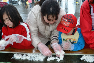 子ども二人と大人の女性がテーブルに座り、手袋をはめて海苔の上にご飯を広げながら海苔巻きづくりをしている様子の写真