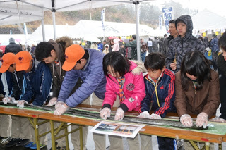 テーブルの上に広げられた長い海苔を前に、参加者の皆さんが手袋を着けて一斉に海苔を巻いている様子が写っている写真