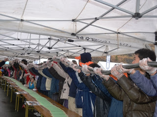 多くの人々がテントの下で並び、完成した長い海苔巻き協力して持ち上げている様子が写っている写真