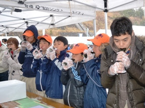 テントの下で数人が並んで座り、それぞれが手に持った海苔巻きを食べている様子が写っている写真