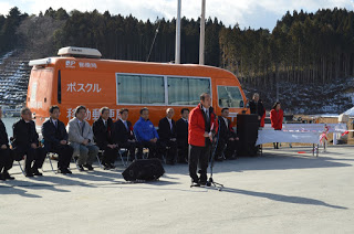 ポスクルと書かれたオレンジ色の大型ワゴン車の前に一列に並んだ椅子に関係者が座り、その前で赤い法被を着た男性が、スタンドマイクに向かって話をしている様子の写真