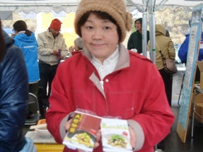 屋外のテントの下で、赤い上着と茶色い帽子を着た女性が「めかぶ納豆」と書かれたパッケージを両手に持ち、カメラに向かって見せている写真