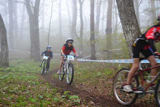 霧の立ち込める森林の中で、ゼッケンをつけた子どもの選手たちが細い土の道をマウンテンバイクで走り抜けている写真