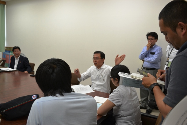 白いワイシャツの男性が会議室のテーブルで手を大きく動かしながら発言しており、それを囲んで数名の記者が真剣な表情でメモや録音をしている写真