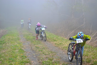濃い霧の中、未舗装の山道をゼッケンをつけた選手たちがマウンテンバイクを押しながら歩いている写真