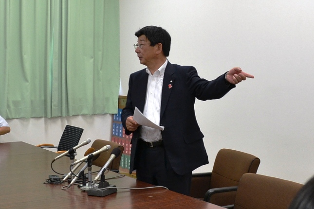 数本のマイクが置かれている机の前に佐藤町長が立ち、資料をもって何かを指し示しながら話をしている様子の写真