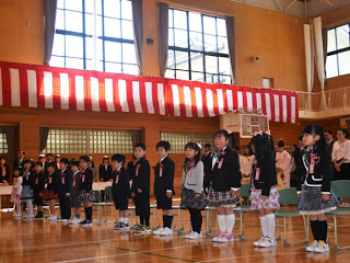 紅白幕が掲げられた体育館で新入生の子どもたちが整列して立ち、後ろには保護者が見守っている入学式の写真。
