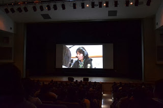 暗くなったホールのスクリーンにインタビューを受けている女性の映像が映し出されている様子を写した写真