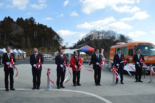 屋外で赤い法被を着た男性を中心に7名の男性が横一列に並び、テープカットを行っている写真