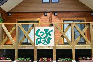 木造の建物の外壁に「くつろぐはうす」と緑色で書かれた看板が掲げられ、周囲のプランターには色とりどりの花が植えられている建物外観の写真