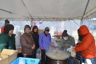 テントの下の大きな鍋から湯気が上がり、その周りに7人の女性が集まっている写真