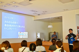 資料が映し出されたスクリーンの右側に立つ講師の話を、椅子に座っている受講者が熱心に聞いている様子を後方から撮影した写真