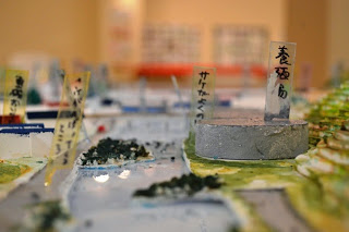 グレー色の円柱型に「養殖場」と手書きで書かれたラベルが貼られ、周囲に森のような模型が並ぶ、港周辺の立体模型の接写写真