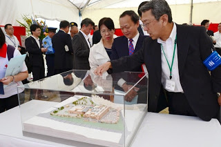 スーツ姿の男性が透明なケース越しに病院施設の模型を指さして説明し、周囲の来賓とともに見学している写真