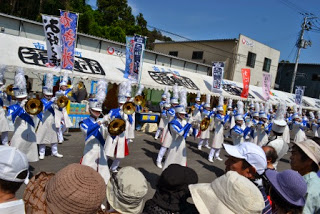 後方にテントが立ち並び、多くの観客が見ている通路で、お揃いの白い衣装を身にまとい、それぞれ楽器を持ったマーチングバンドのメンバーが間隔をあけて立ち演奏を行っている写真