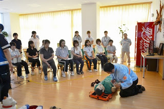 太陽の光が反射する明るい室内に座っている受講者たちの前で、講師が人形を使って心肺蘇生法の実演を行っている様子の写真