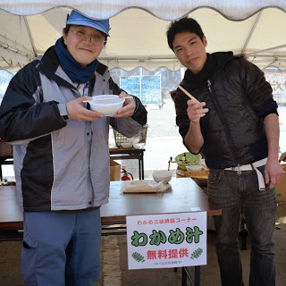 屋外イベントのテントの下で、2人の男性が「わかめ汁無料提供」と書かれたポップの前に立ち、右側の男性は右手に割りばしを持ち、左側の男性は手にスープの入った器を持っている様子の写真