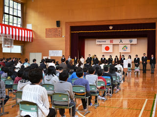 体育館の舞台に「祝 入学」と書かれた横断幕と日の丸の旗が掲げられ、前方に先生方が横一列に並び、在校生と新入生が椅子に座って前方を見ている入学式の様子の写真