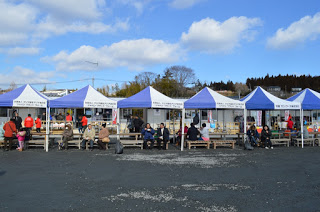 青と白の配色のテントが並び、下に置かれたベンチに人々が座っている様子の写真