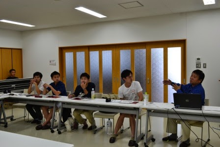 青い服の男性がパソコンが置かれた席に座って話しているのを、五人の若者が真剣な表情で聞いている教室内の写真