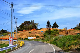 舗装された道路のカーブの先で、山を切り崩して整地が始まっており、ショベルカーなどの重機が土の斜面で作業をしている写真
