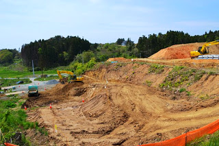 緑に囲まれた中で、土の高台の上や斜面でショベルカーやトラックが造成の作業をしている写真