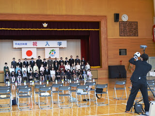 体育館の舞台に新入生と先生、保護者の皆さんが並んで座り、カメラマンが記念写真を撮影している入学式の集合記念撮影の写真。