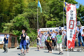 「伊里前福幸商店街」と書かれたのぼり旗の前で、「伊里前福幸商店街」を一文字ずつ持ってステージに立つ人々と、前列に立つスタッフの方々が手を振りながら踊っている様子の写真