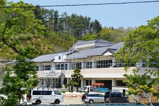 白とベージュの鉄筋コンクリート造で2階建ての校舎に足場が設置され、校庭に複数の工事関係車両が停まっている小学校の外観写真