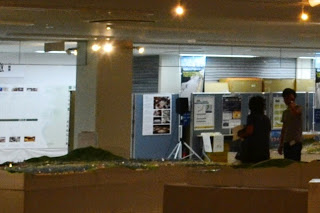 周囲には地形模型や説明パネルが並べられていて、展示資料が掲示されたパネルの前に二人の人物が並んで立っている展示会場の写真