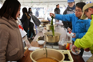 冬の寒さの中、テントの下で人々がカセットコンロと鍋が設置されたテーブルを囲み、鍋の中のお湯に緑色のワカメをくぐらせている様子の写真