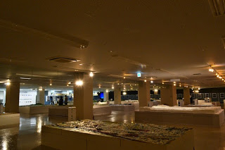 薄暗い照明の広い室内に、複数の白い台の上に模型が並べられており、奥にはパネル展示も見える写真