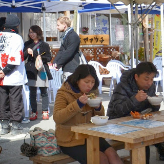 木製のテーブルとベンチが設置された屋外の青と白のテントの飲食スペースで、木製のテーブルに座った2人が温かい汁物が入った器を持ち食事をしているイベント会場の写真