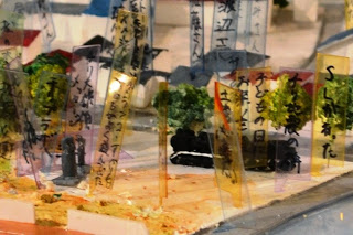 木やラベルが立ち並ぶ町中に黒い小型列車模型が置かれている情景の拡大写真