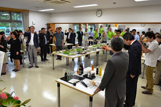 広い会場に多くの来場者が集まり、立食形式で料理が並ぶテーブルを囲みながら乾杯している様子の写真