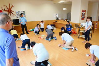 講師たちが周囲で見守るなか、広めの部屋で受講者が胸骨圧迫の実習を行っている様子の写真