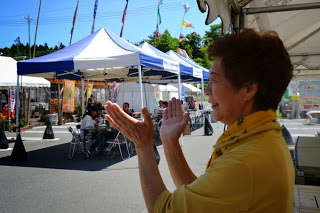 テント下にいる黄色い服を着た女性が、手をたたいて楽しそうに笑いながらダンスを見守っている写真